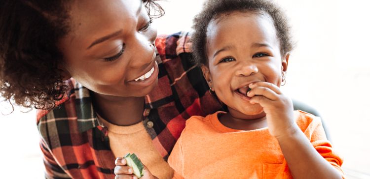 A black mother holds her baby girl who smiles at the camera with one hand in her mouth and a cucumber slice in the other