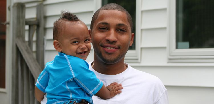 A black man holds his smiling baby boy who has a curly mini-mohawk hairstyle