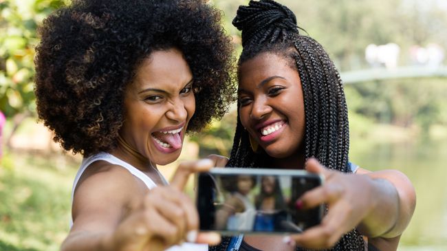 A woman with an afro sticks her tongue out as her friend in braids smiles demurely as they take a selfie together.